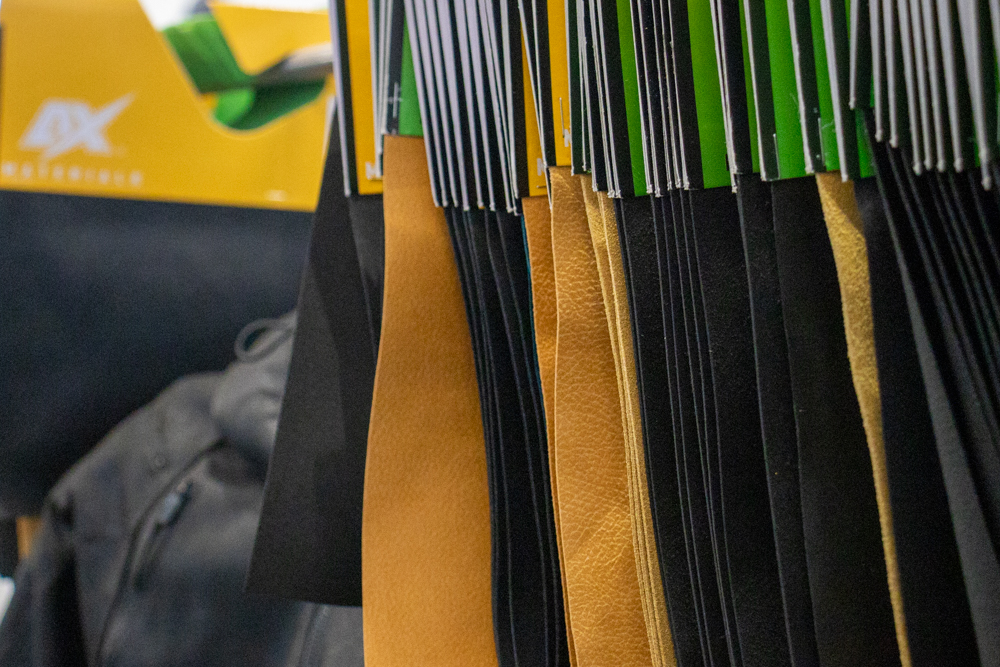 Close-up of glove material samples on hangers showing textures and colors from black to tan, with AX® Materials branding in the background.